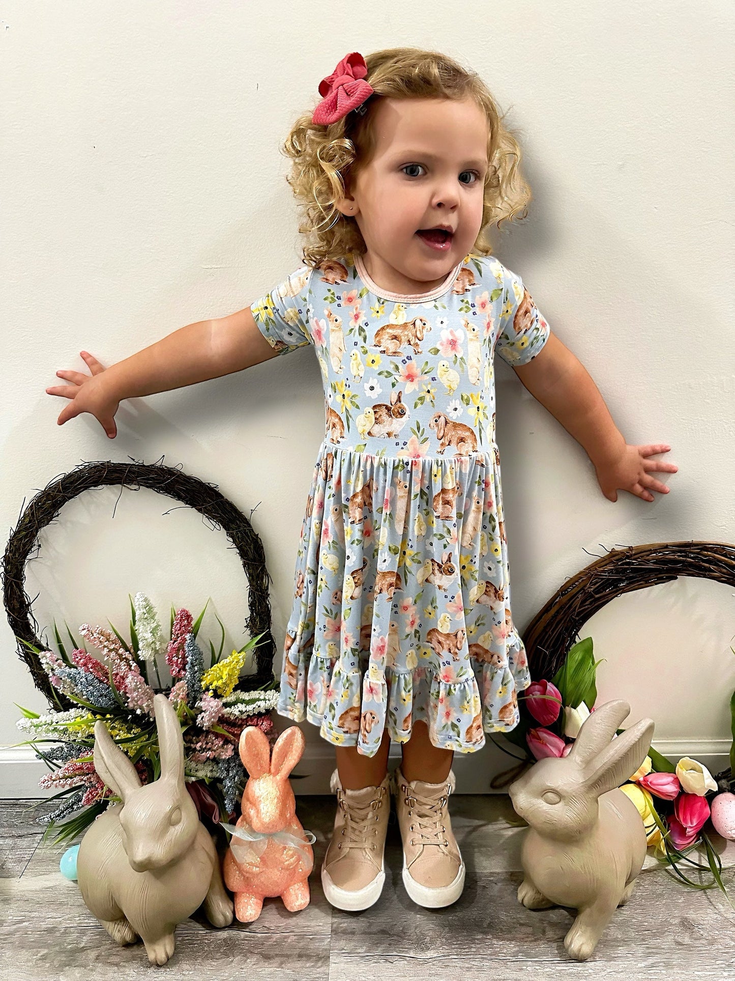 Toddler wearing Bunny Blooms Twirl Dress with floral and animal print, featuring ruffles and bow; surrounded by rabbit statues and spring flowers.