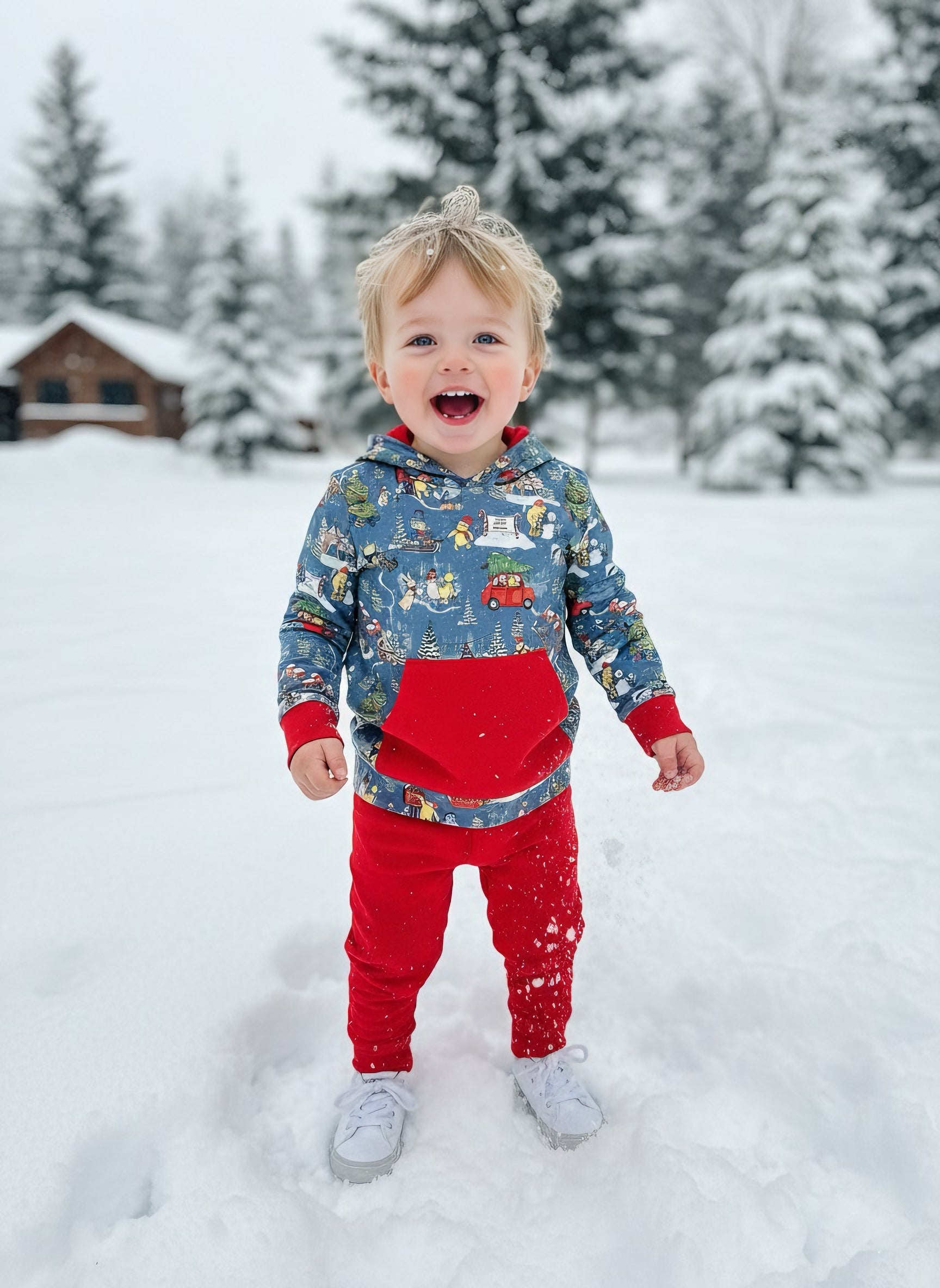 Child in a blue and red outfit standing in the snow with trees and a cabin in the background