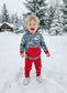 Child in a blue and red outfit standing in the snow with trees and a cabin in the background