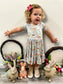 Toddler wearing Bunny Blooms Twirl Dress with floral and animal print, featuring ruffles and bow; surrounded by rabbit statues and spring flowers.
